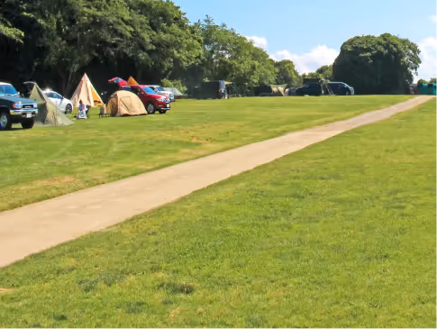 Grassy campsite with tents, parked cars, and a paved path leading into a tree-lined area under a partly cloudy sky.