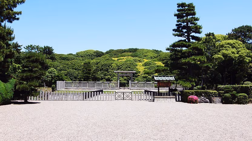 Mozu Tombs surrounded by dense green trees under clear blue sky, viewed from a gravel path with a wooden fence and torii gate.