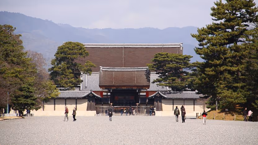 Wide gravel pathway leading to the entrance of a traditional Japanese palace building surrounded by green trees and distant mountains.