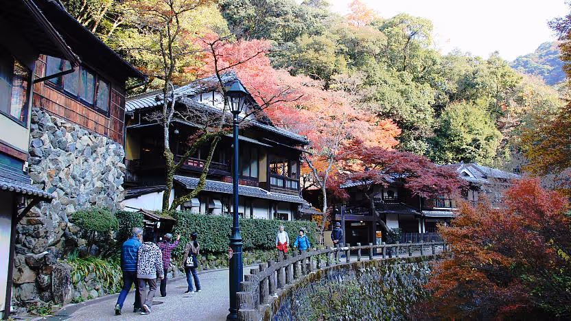 People walking along a curved stone railing path by traditional Japanese houses with autumn trees in the background.