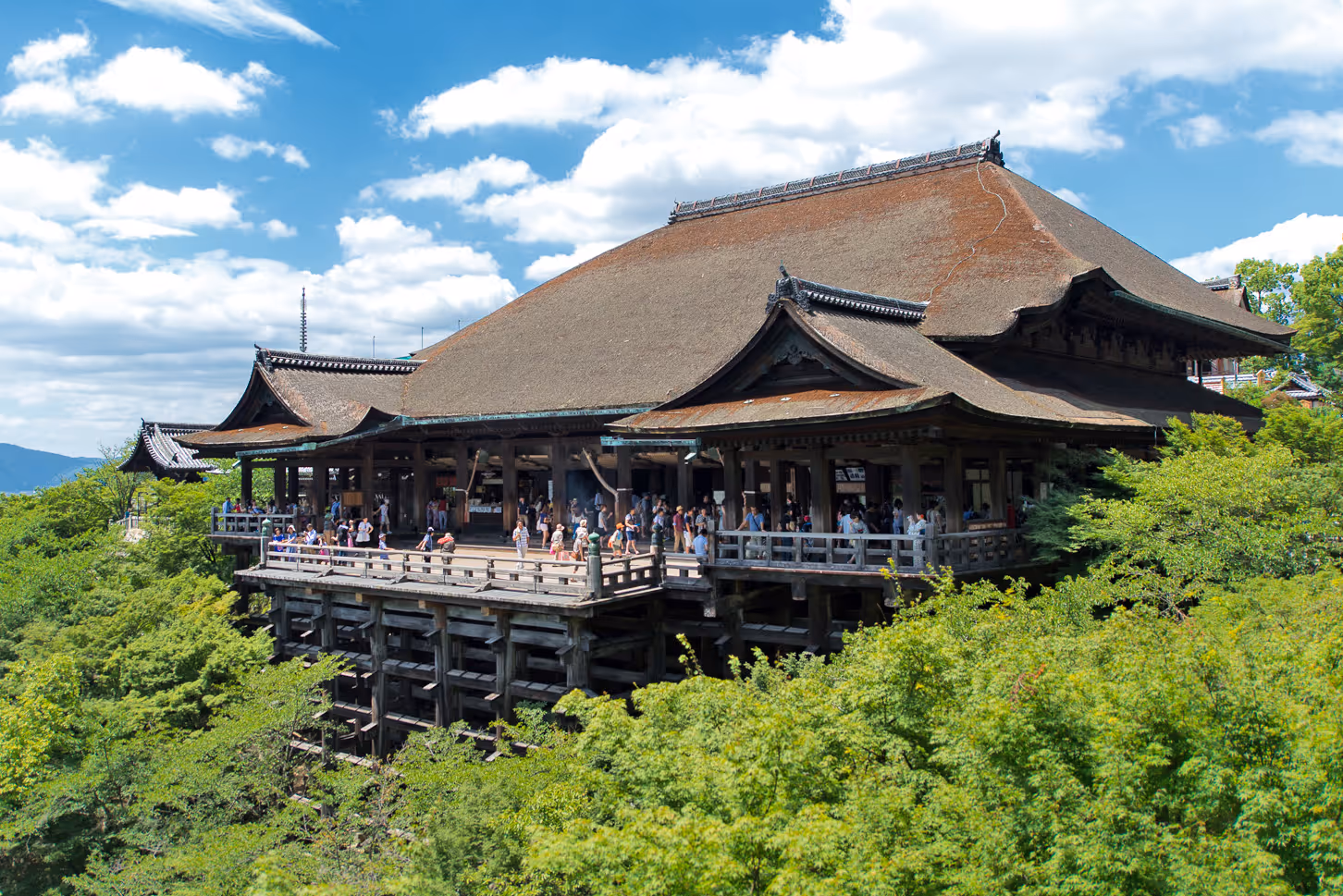 Kiyomizu-dera Buddhist temple in Kyoto surrounded by green trees under a blue sky with clouds.