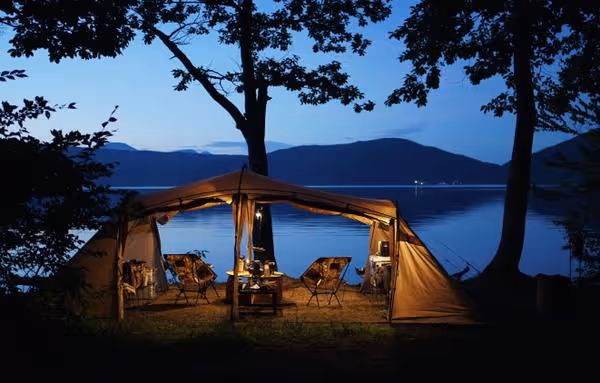 Illuminated camping tent with chairs and table set inside by a calm lake at dusk with silhouetted trees and distant mountains.