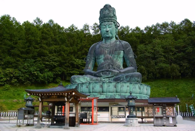 Large green Buddha statue seated in meditation in front of traditional wooden structures with dense green trees in the background.