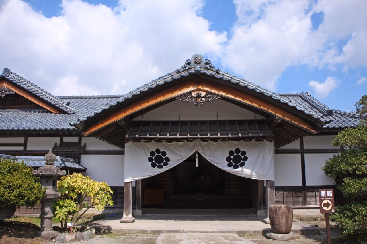 Traditional Japanese building entrance with a tiled roof, white fabric drapes featuring black circular patterns, stone lantern, and greenery under a partly cloudy sky.