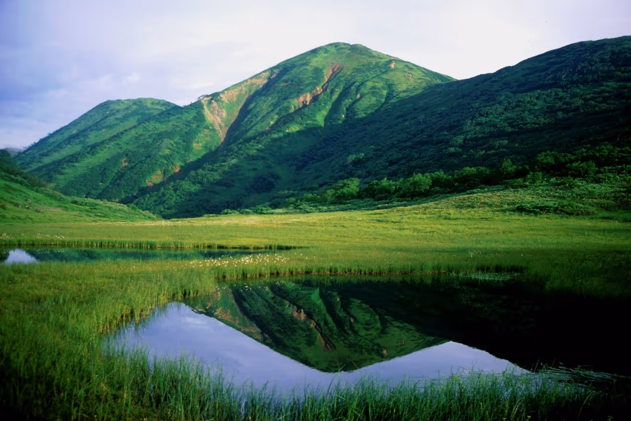 Green mountains reflected in a calm lake surrounded by grassy fields under a cloudy sky.