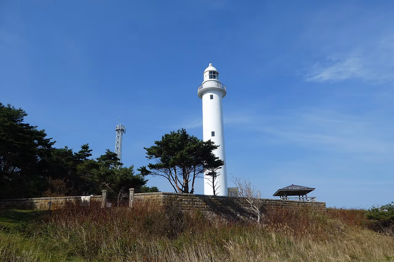 Tall white lighthouse on a grassy hill with trees and a small pavilion under a clear blue sky.
