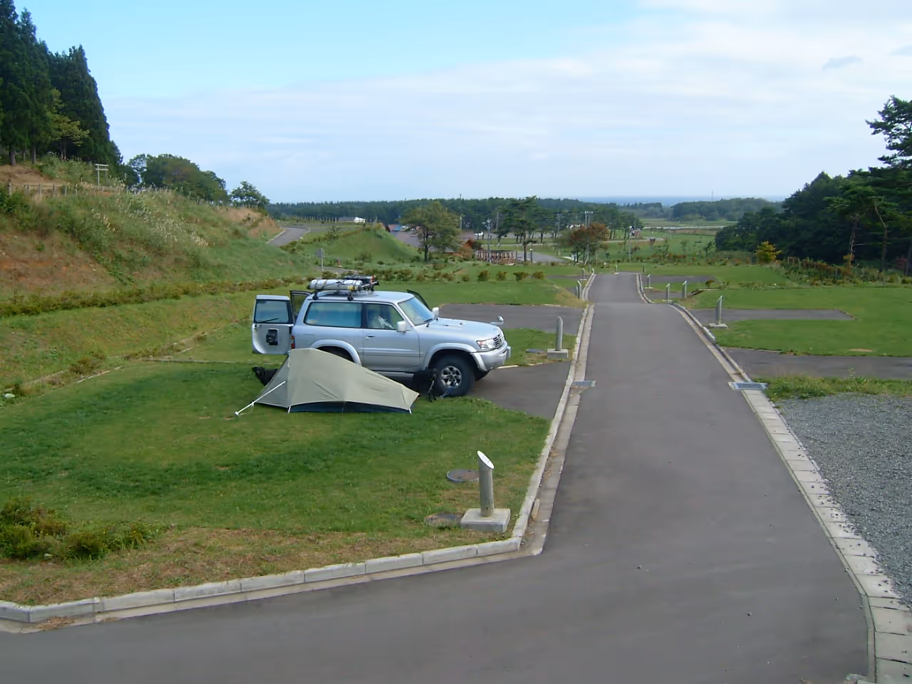Silver SUV parked next to a green tent on grassy area beside a paved road at a campsite.