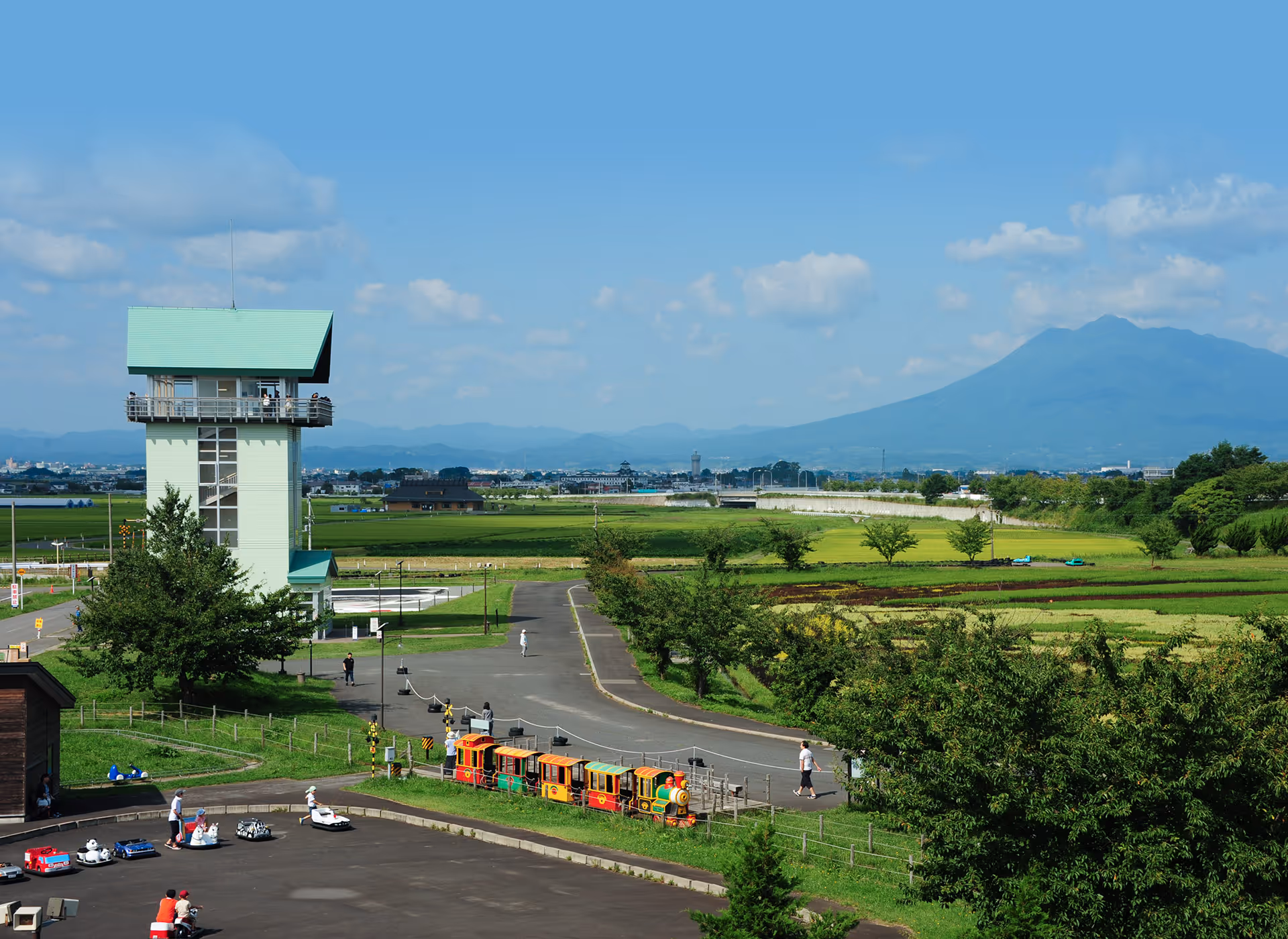 A green-roofed observation tower overlooking a park with a small colorful train and people riding pedal cars, with green fields and a mountain in the background under a blue sky.