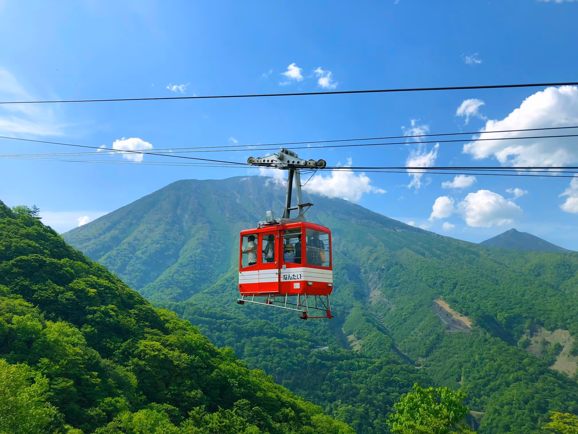 Red and white ropeway cable car with passengers traveling over green forested mountains under a blue sky with scattered clouds.