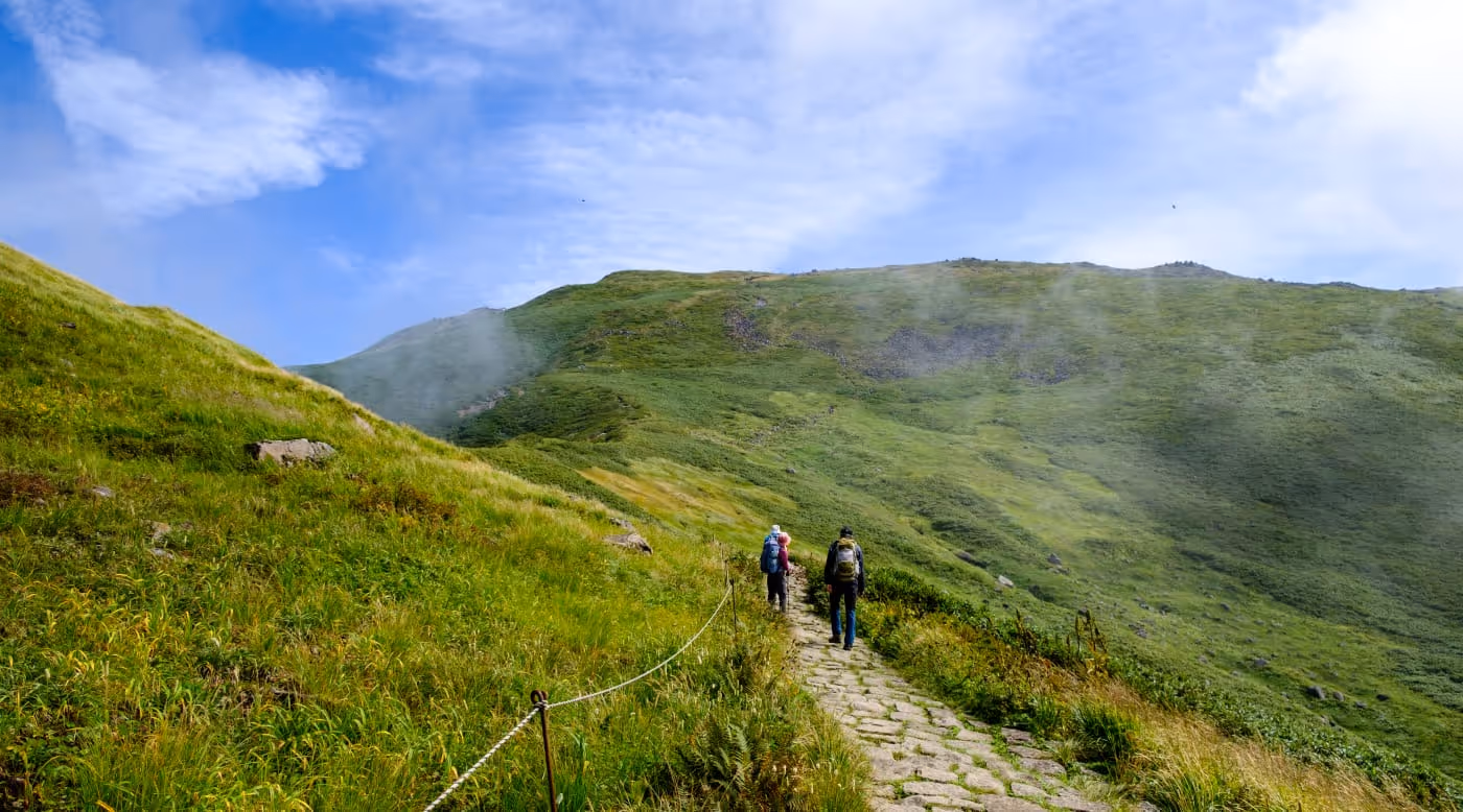 Two hikers walking on a stone path through green grassy hills under a blue sky with some clouds.
