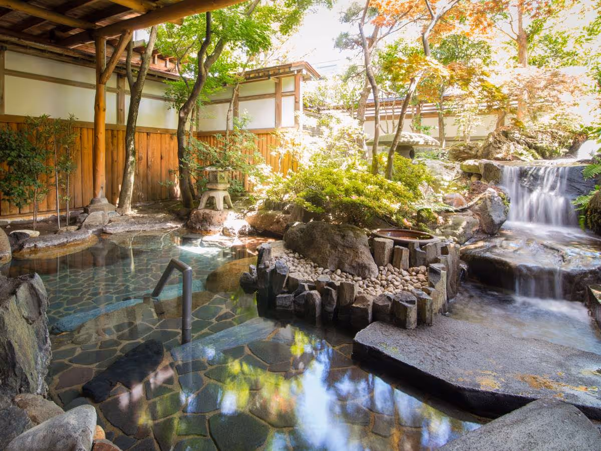 Outdoor Japanese onsen with clear water, stone floor, surrounding greenery, wooden fence, and a small waterfall.