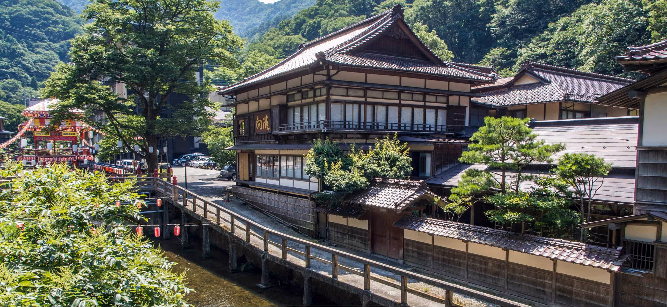 Traditional Japanese wooden building by a river with a wooden bridge and lantern decorations, surrounded by lush green trees and mountains.