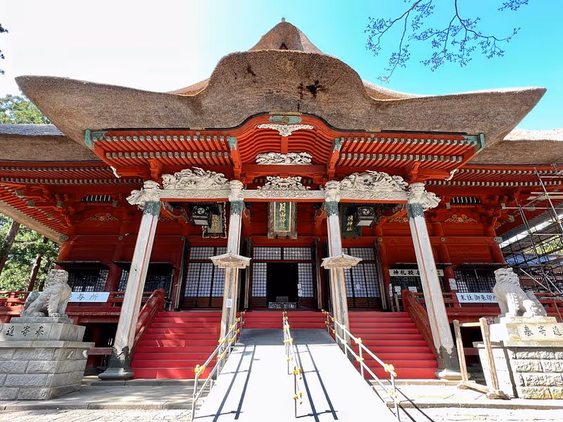 Front view of a traditional Japanese temple with a large thatched roof, red wooden pillars, and stone lion statues at the entrance.