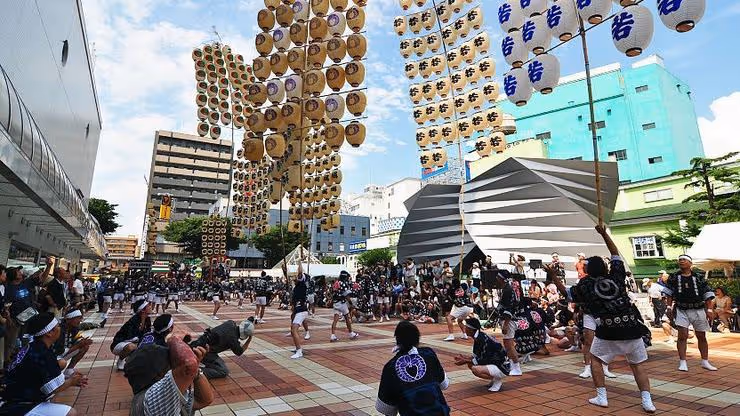 Performers balancing large wooden poles adorned with many paper lanterns during the Kanto Matsuri festival in an urban plaza.