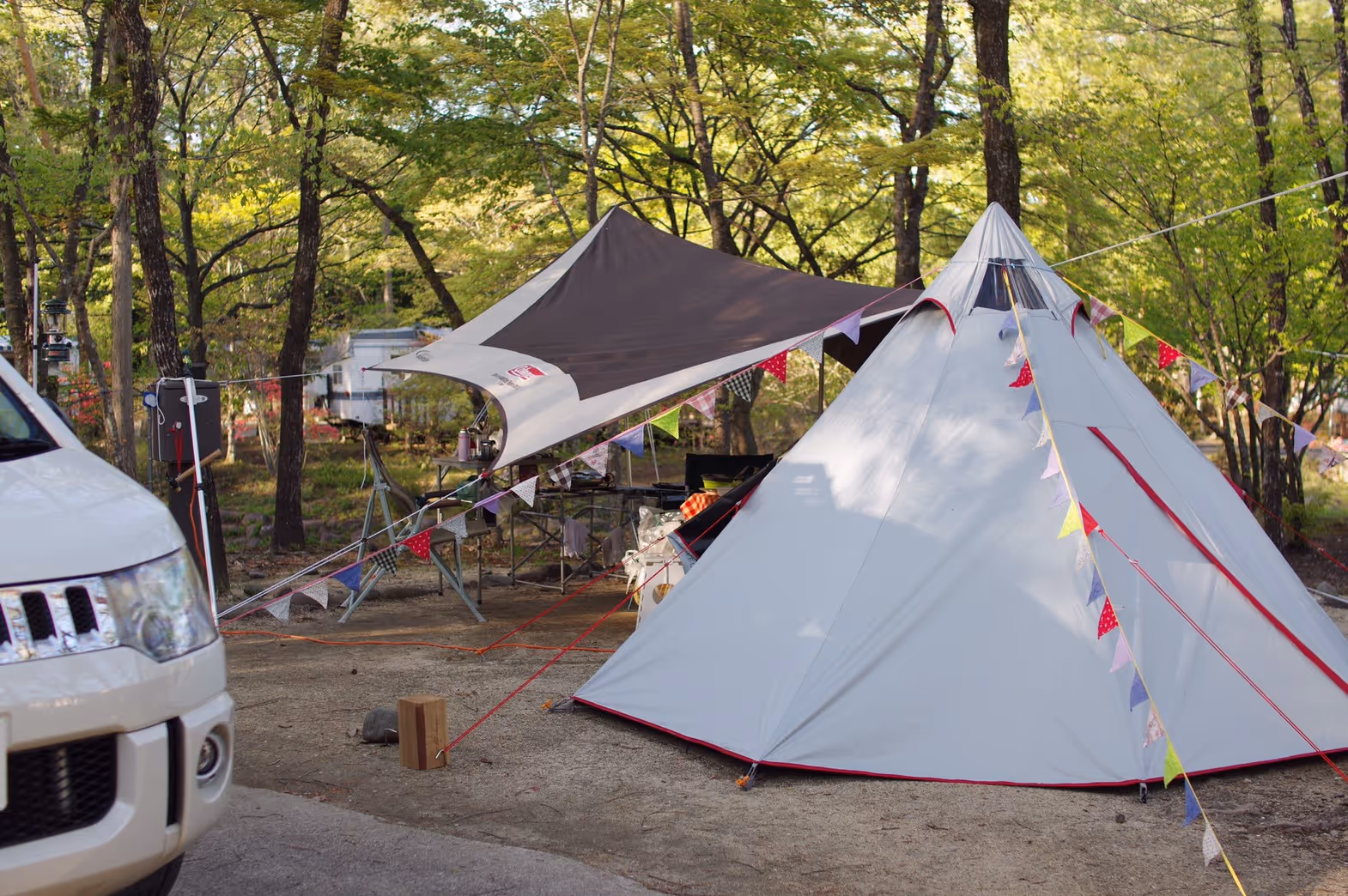 White teepee-style tent decorated with colorful triangular flags under a brown canopy in a wooded campsite with a white vehicle partially visible on the left.