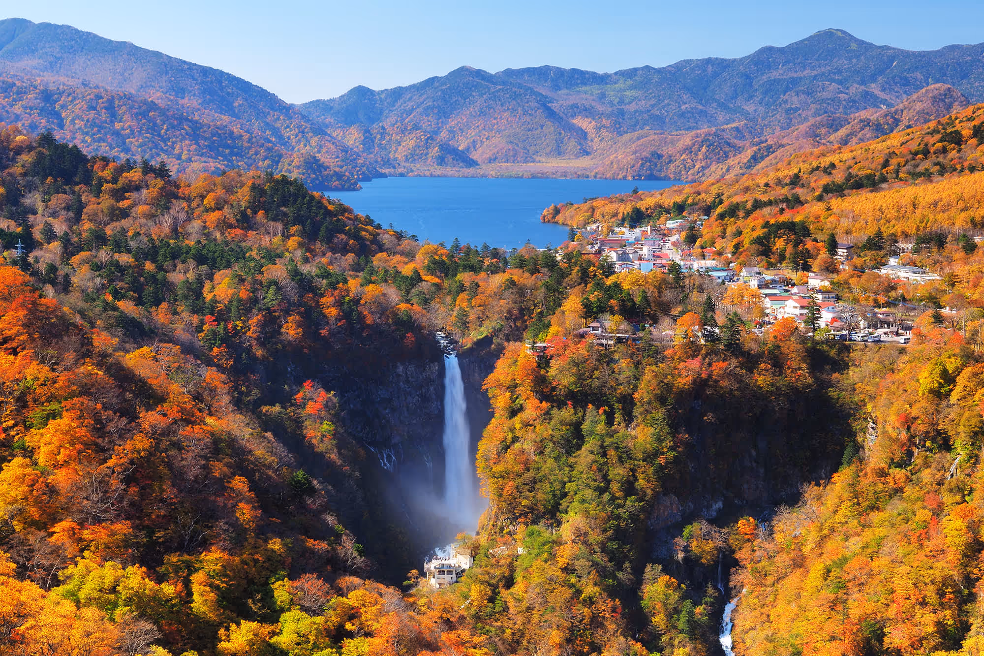 A waterfall surrounded by colorful autumn trees with a blue lake and mountains in the background.