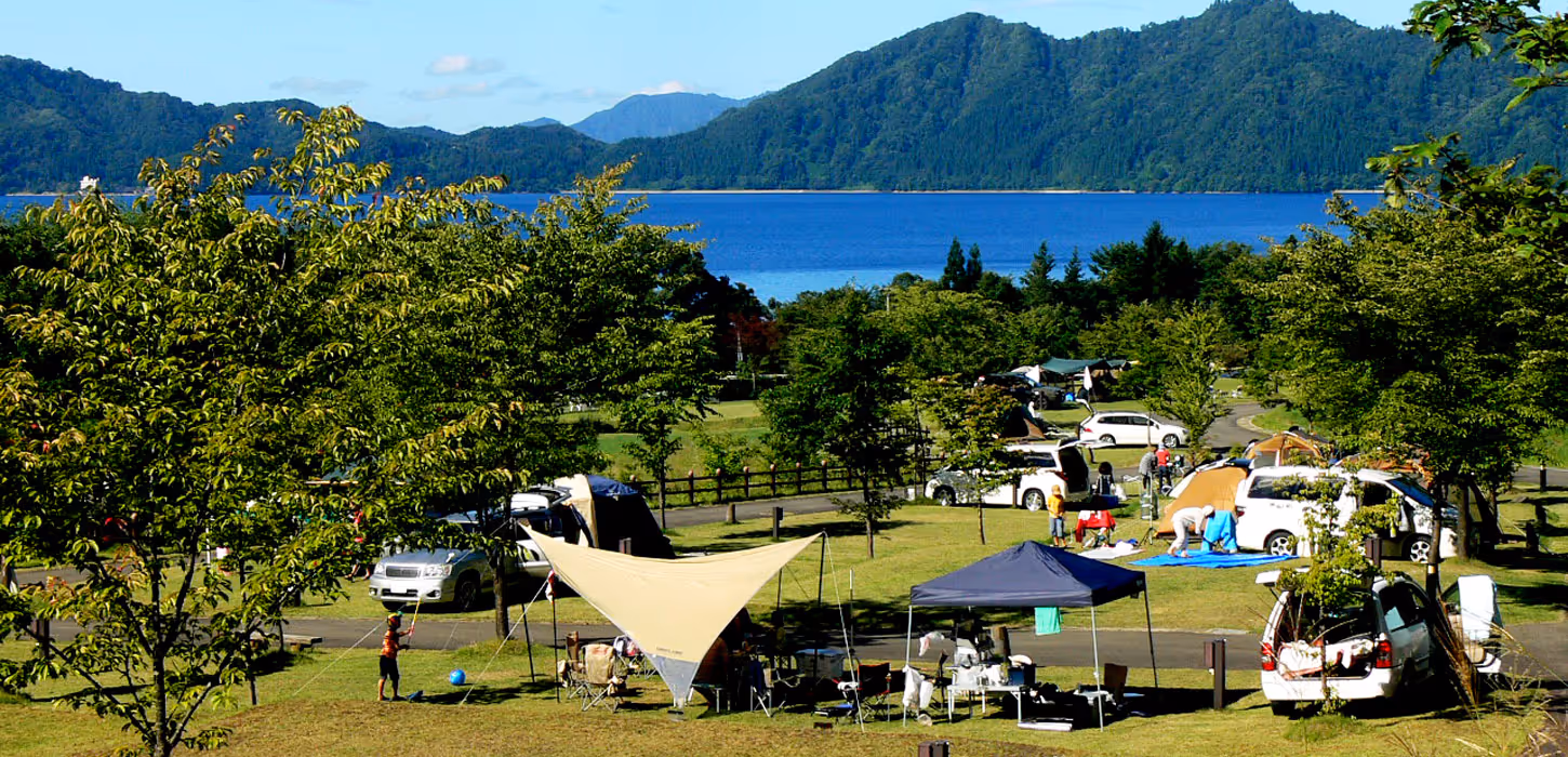 Campsite with tents, vehicles, and people setting up camping gear near a lake surrounded by green hills under a clear blue sky.