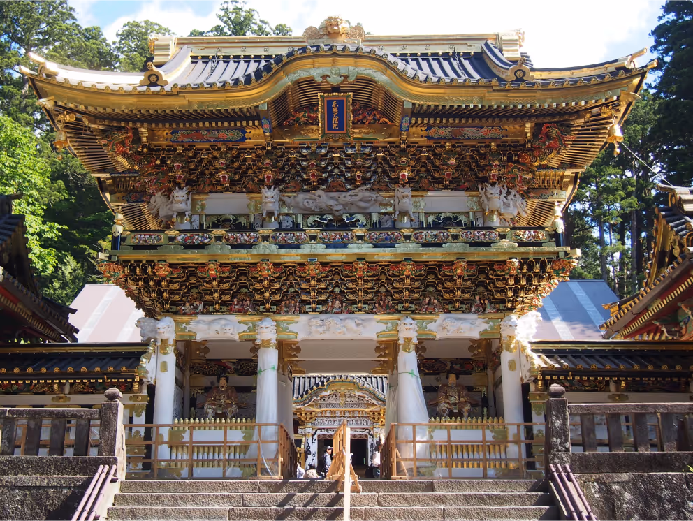Ornate golden and white traditional Japanese temple gate surrounded by trees and stone steps.
