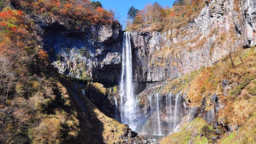 Tall waterfall cascading down rocky cliffs surrounded by autumn-colored trees under a clear blue sky.