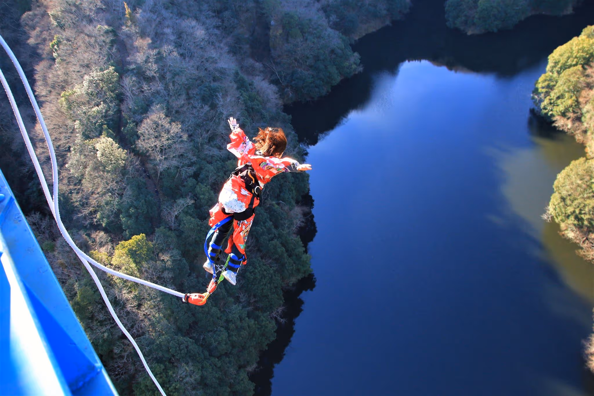 Person in a red outfit bungee jumping over a forested river from a blue platform.