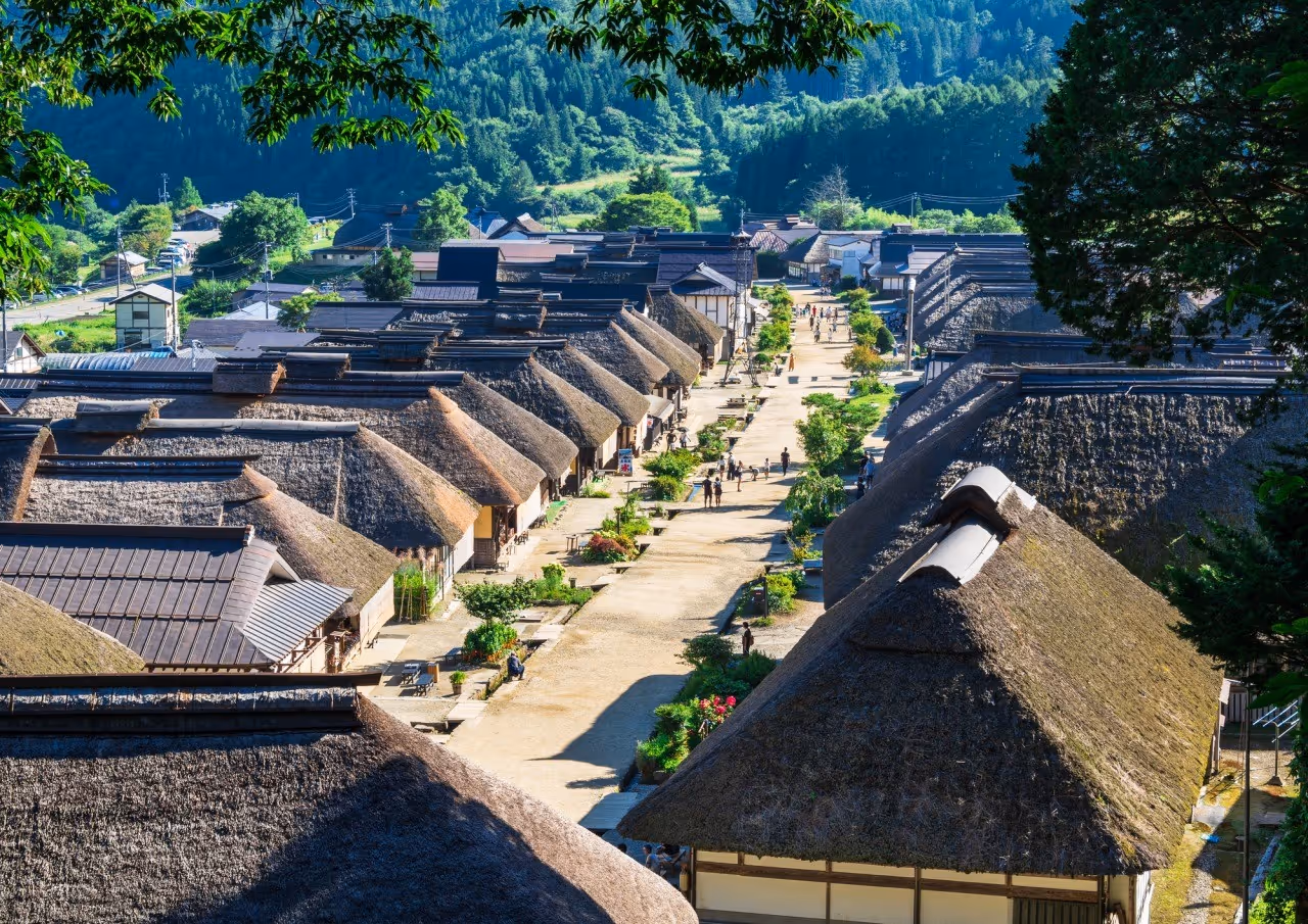 A traditional Japanese village with thatched roof houses lining a sunlit pathway surrounded by green trees and forested hills.