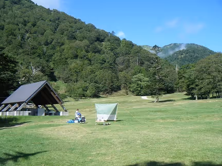 Campground with a grassy field, small tent, shelter structure, and person among green forested hills under a clear blue sky.