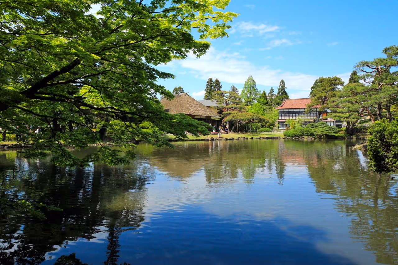 Serene Japanese garden with traditional buildings, lush green trees, and a calm reflective pond under a blue sky.
