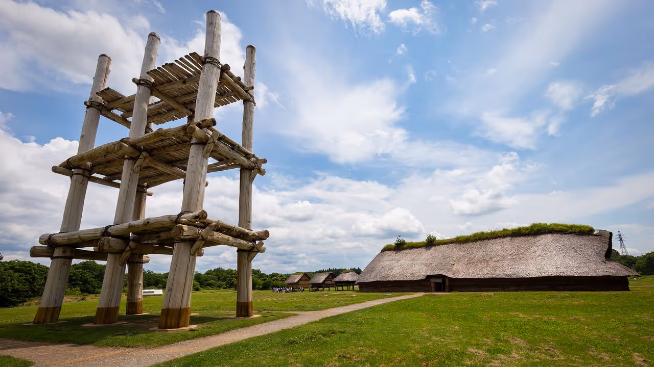Large wooden reconstruction of a Jomon period watchtower next to traditional thatched-roof buildings under a partly cloudy sky.