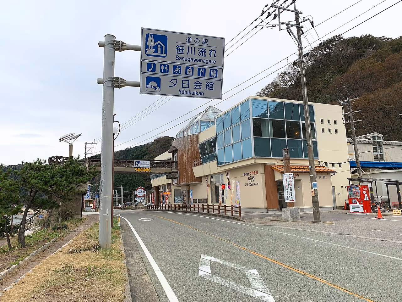 Roadside view of a modern building complex with signage indicating Sasagawanagare and Yuhikaikan, featuring a vending machine and power poles.