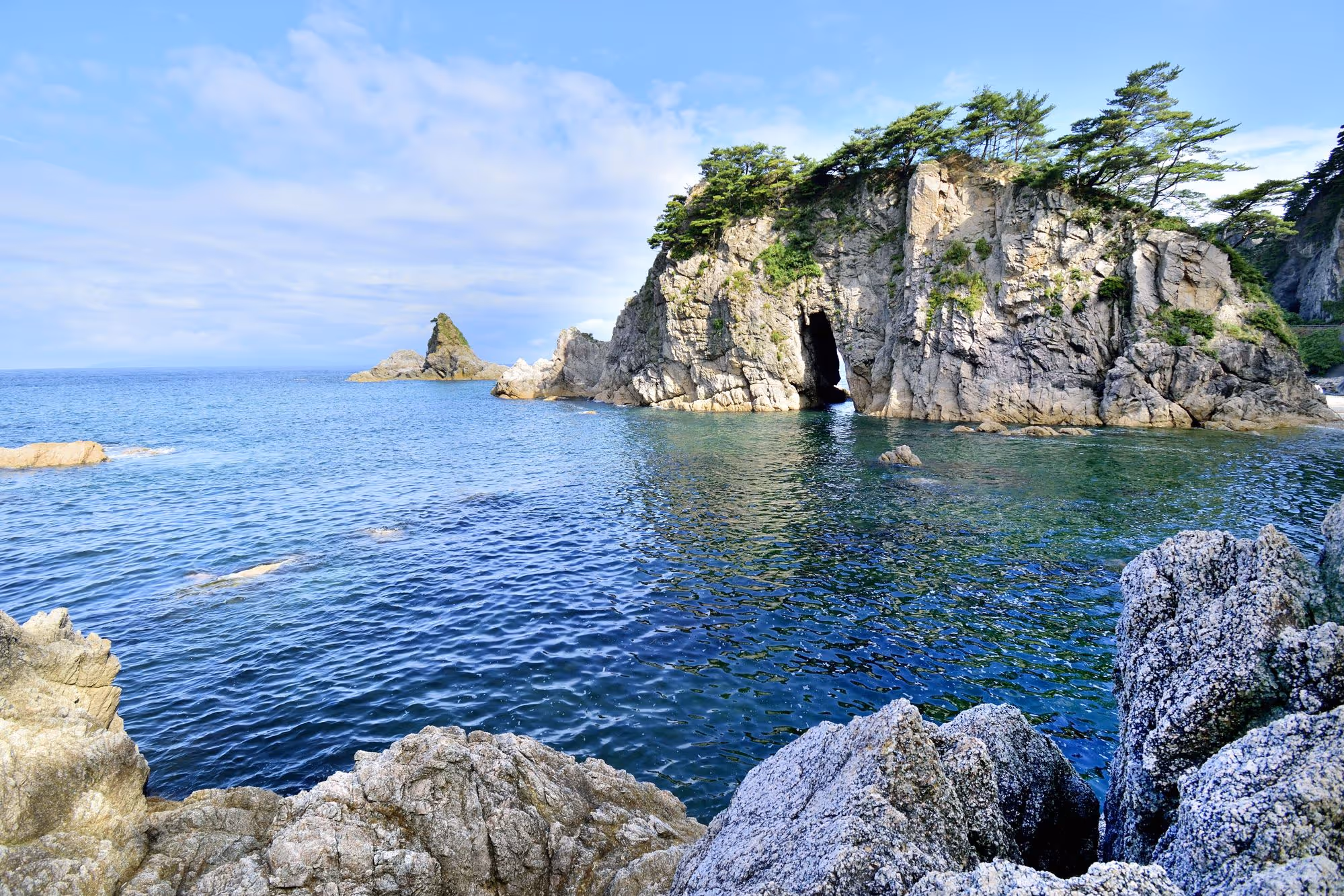 Rocky coastal cliffs with pine trees and a sea cave opening, overlooking calm blue ocean waters under a partly cloudy sky.