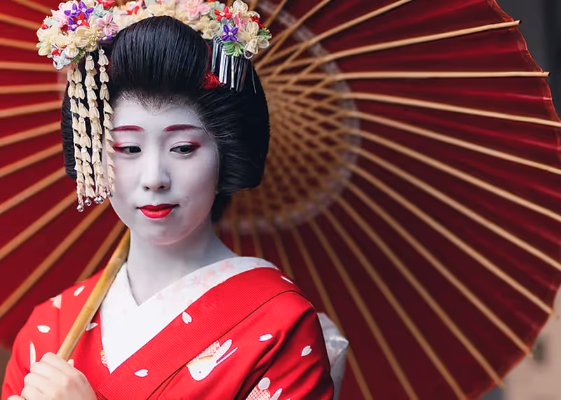 Woman dressed as a maiko with traditional white makeup, floral hair ornaments, and a red kimono holding a red umbrella.