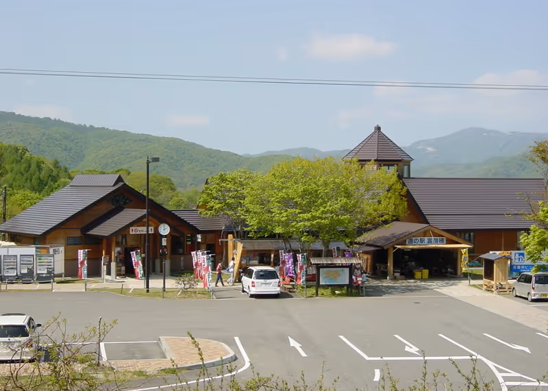 Roadside station with brown-roofed buildings surrounded by green trees and mountains in the background on a clear day.