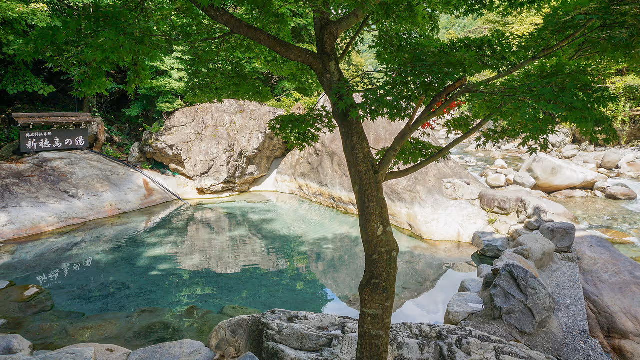 Outdoor natural hot spring pool surrounded by large rocks and green trees with clear water reflecting the surroundings.