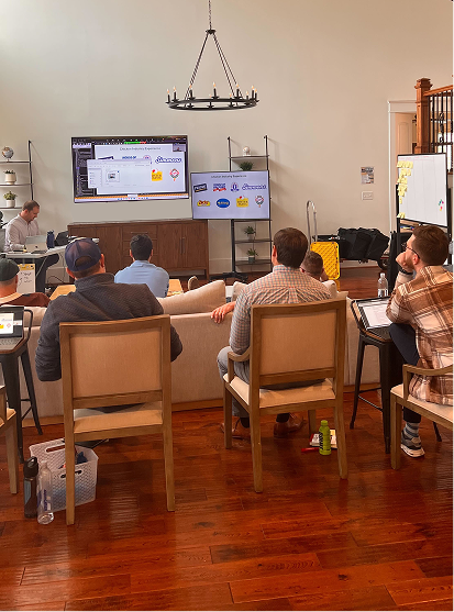 Group of five people seated in a room watching two large screens displaying logos and graphics during a presentation.