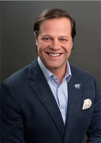Smiling man wearing a navy blazer with a white pocket square and a light blue checkered shirt against a gray background.
