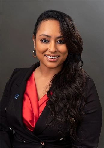 Smiling woman with long wavy hair wearing a black blazer and red blouse against a dark background.