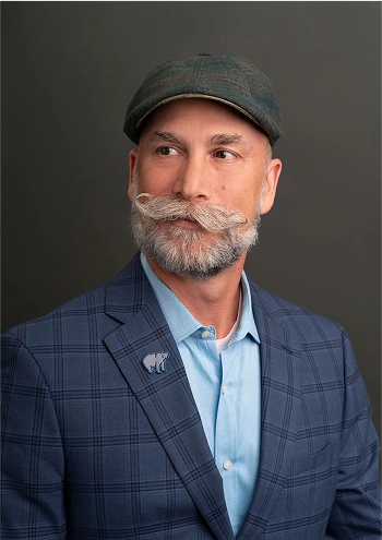 Man with gray styled mustache and beard wearing a green flat cap, blue plaid blazer, and light blue shirt against a dark background.