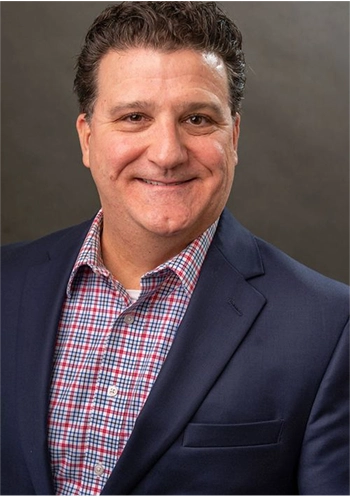 Smiling man wearing a dark blazer and red checkered shirt against a plain background.
