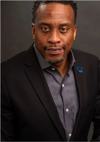 Portrait of a man wearing a black blazer over a gray shirt with a serious expression against a dark background.
