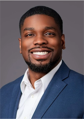 Smiling man with short hair and a beard wearing a blue blazer and white shirt against a gray background.