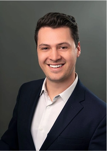 Smiling young man with dark hair wearing a white shirt and dark blazer against a gray background.
