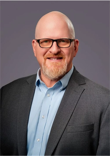 Smiling bald man with glasses and a red beard wearing a gray suit jacket and light blue shirt against a plain gray background.
