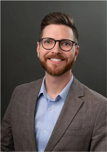Smiling man with glasses, short brown hair, and beard wearing a light blue shirt and brown checkered blazer against a dark background.