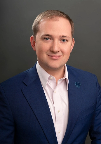 Portrait of a smiling man in a blue suit jacket and light pink shirt against a plain dark background.