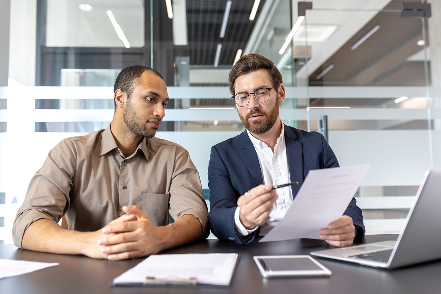Two men sitting at a desk in an office, one in a blue suit and glasses reviewing a document while the other listens attentively.