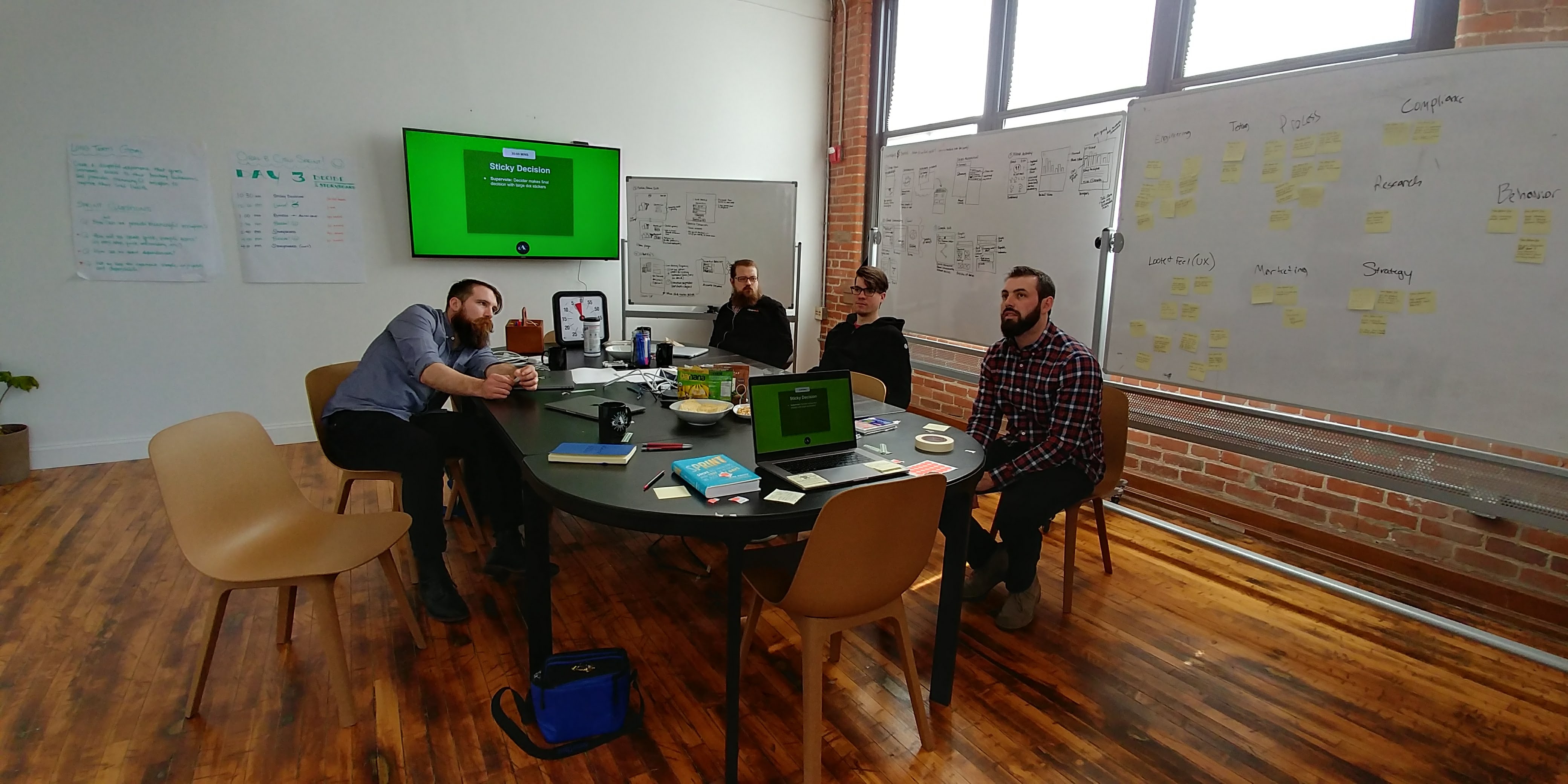 A picture of three men sitting in a conference room in front of a whiteboard near a TV with lots of design sprint-related sketches and notes