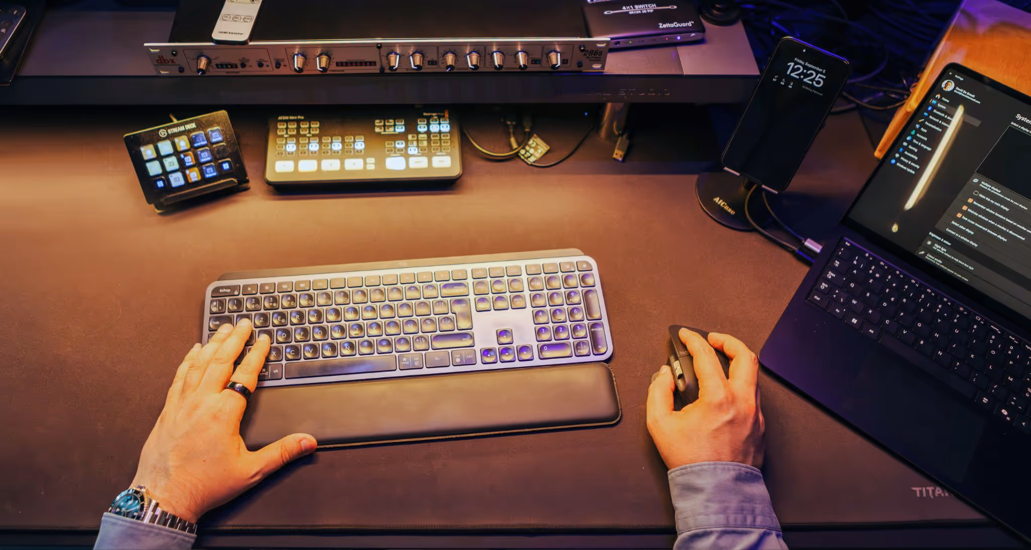 Person using a wireless keyboard and mouse on a desk with a laptop, smartphone on stand, and audio equipment.