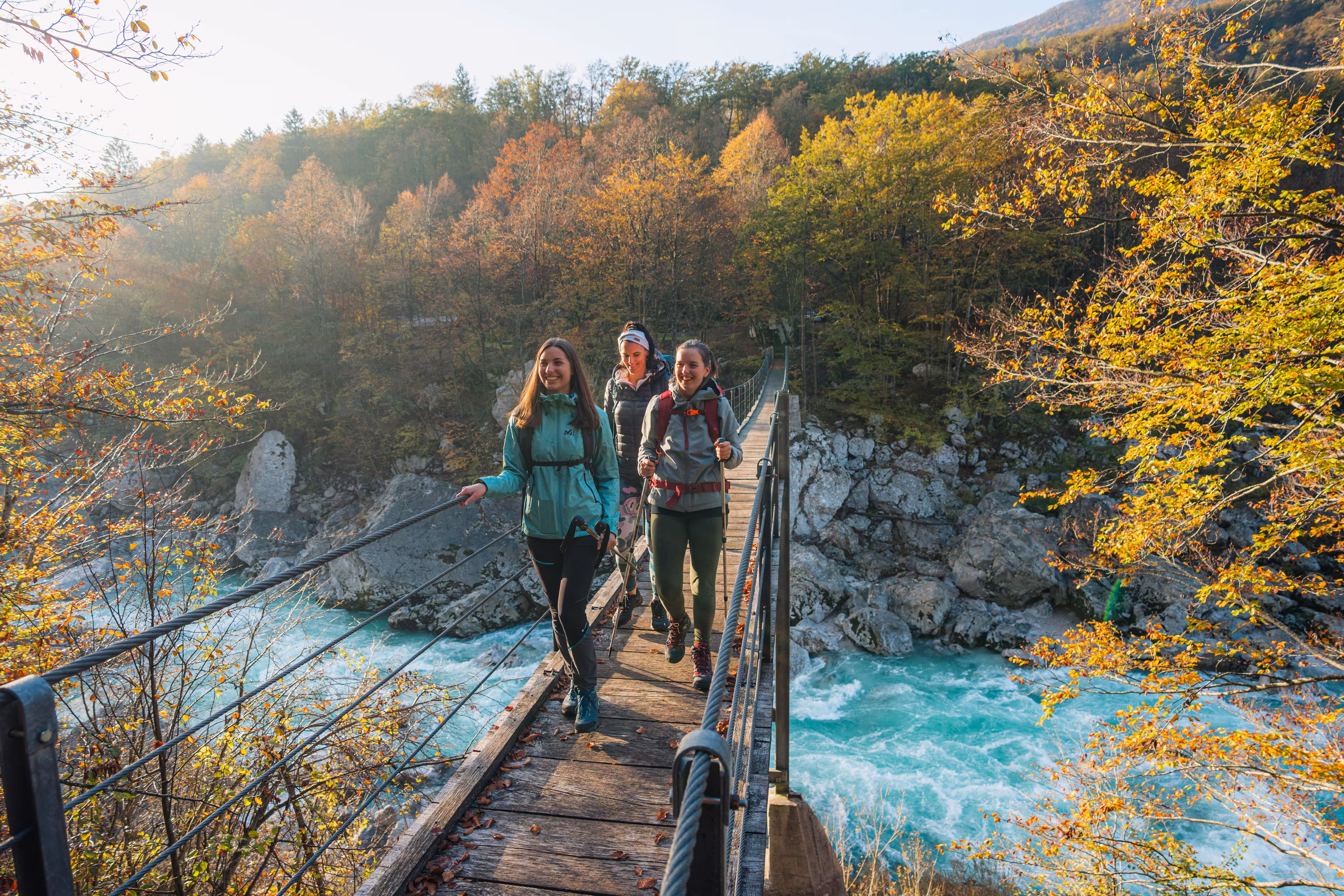 Drie wandelaars steken een hangbrug over de Soča over, tussen herfstkleurige bossen