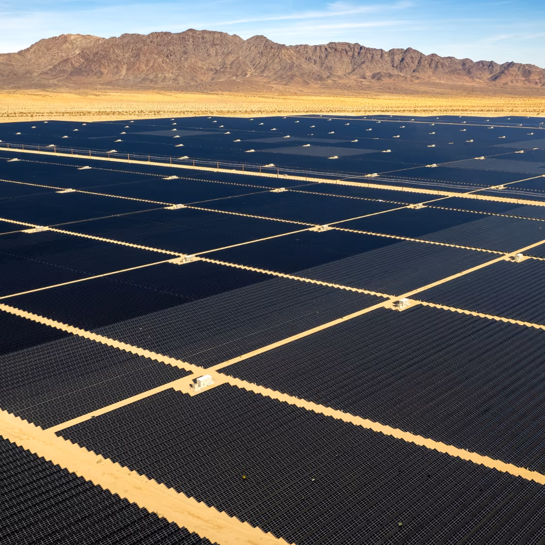 A vast solar farm with rows of solar panels in a desert landscape with mountains in the background under a clear blue sky.