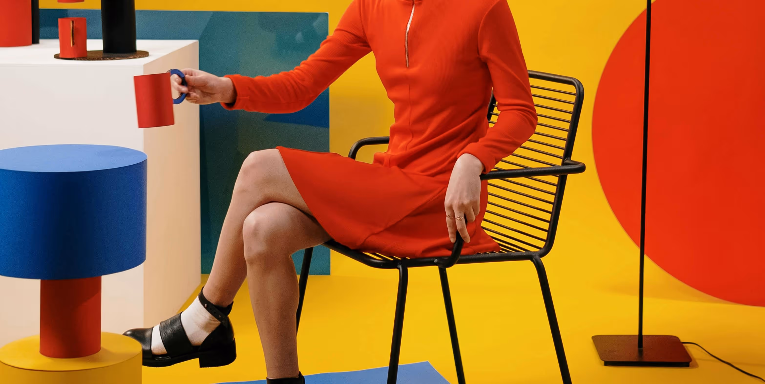 Person in a red dress sitting on a black wire chair holding a red mug in a colorful room with yellow, blue, and red geometric shapes.